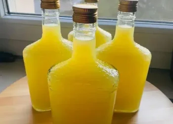 Four square glass bottles filled with bright yellow liquid, crafted from a traditional limoncello recipe and sealed with gold screw caps, are arranged on a wooden surface near a window.