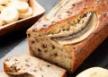 A loaf of angular banana bread with chocolate chips is sliced on a dark surface. A bowl of banana slices and a whole banana are beside the bread.