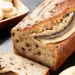 A loaf of angular banana bread with chocolate chips is sliced on a dark surface. A bowl of banana slices and a whole banana are beside the bread.