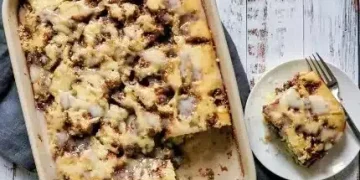 A baked cinnamon crumb cake with icing, perfect for fans of Cinnamon Recipes, sits in a rectangular dish. One square piece is served on a plate with a fork, all arranged on a rustic white wooden surface.