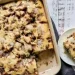 A baked cinnamon crumb cake with icing, perfect for fans of Cinnamon Recipes, sits in a rectangular dish. One square piece is served on a plate with a fork, all arranged on a rustic white wooden surface.