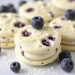 Round blueberry cream cookies are stacked on a white surface, with fresh blueberries scattered around and on top. These white chocolate blueberry Oreos have visible blueberry pieces and a creamy texture.