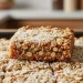 A close-up of a chewy oat bar topped with shredded coconut, resting on a tray of similar bars. The golden-brown color and visible whole oats evoke a classic homemade feel—perfect for anyone seeking an authentic Anzac slice recipe.