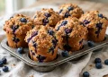 Tall blueberry muffins with golden cinnamon streusel topping and bursting berries in a muffin tin
