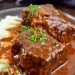 Boneless Beef Short Ribs braised and covered in rich brown gravy, garnished with chopped parsley, served alongside a portion of creamy mashed potatoes on a dark plate.
