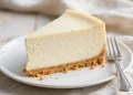 A tall slice of classic New York cheesecake with graham cracker crust on a white plate