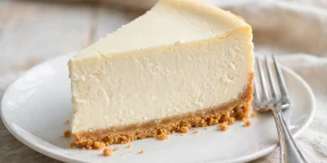 A tall slice of classic New York cheesecake with graham cracker crust on a white plate