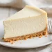 A tall slice of classic New York cheesecake with graham cracker crust on a white plate