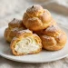 Cream puffs filled with whipped cream, dusted with powdered sugar, stacked on a white plate