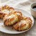 Golden flaky cream cheese danish pastries with vanilla glaze drizzle on a white plate with coffee