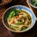 A bowl of noodle soup with green vegetables and herbs, served in a patterned blue and white bowl on a wooden table with chopsticks on the edge, evokes the comforting flavors of a classic phat si-io recipe.