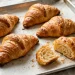 Golden-brown flaky croissants with visible layers on parchment, one broken open showing airy interior