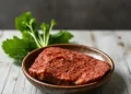 A wooden bowl holds a slice of uncooked, seasoned plant-based meat. Fresh green herbs are placed on the rustic wooden surface in the background, perfect for those exploring free carnivore diet recipes with a modern twist.