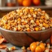 A large wooden bowl filled with an autumn Chex mix recipe—cereal, candy corn, and popcorn—sits on a table decorated with small pumpkins and autumn leaves. The softly blurred background adds to the cozy fall atmosphere.