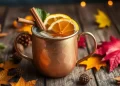 A copper mug filled with an autumn mule drink, garnished with orange slices and cinnamon sticks, sits on a wooden table surrounded by autumn leaves, pinecones, and warm ambient lights.