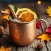 A copper mug filled with an autumn mule drink, garnished with orange slices and cinnamon sticks, sits on a wooden table surrounded by autumn leaves, pinecones, and warm ambient lights.
