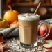 A glass mug of frothy coffee with a cinnamon stick sits on a rustic table, surrounded by apples, pumpkins, autumn leaves, oats, and a cozy knit blanket, capturing the inviting vibe of an apple crisp shaken espresso in autumn.