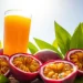 A glass of orange juice is surrounded by whole and halved passion fruits with yellow pulp, resting on green leaves under bright sunlight against a clear blue sky, capturing the essence of refreshing Passion Fruit Juice.