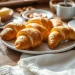 A plate of golden croissants made from a classic gipfeli recipe sits on a wooden table, surrounded by whole almonds, butter, two cups of coffee, and a folded white napkin in a cozy, sunlit setting.