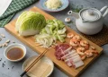 A wooden board with napa cabbage, sliced vegetables, shrimp, and two types of sliced meat—perfect for a Japanese mounjaro recipe. Nearby are dipping sauce, chopsticks, a teapot, garlic, and a small plate of grated ingredients on a gray surface.