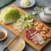 A wooden board with napa cabbage, sliced vegetables, shrimp, and two types of sliced meat—perfect for a Japanese mounjaro recipe. Nearby are dipping sauce, chopsticks, a teapot, garlic, and a small plate of grated ingredients on a gray surface.