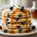Tall stack of fluffy blueberry buttermilk pancakes with maple syrup dripping and fresh blueberries