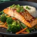 A plate with glazed miso salmon recipe garnished with sesame seeds and herbs, served over sautéed vegetables (broccoli, carrots, snap peas) and a side of white rice.