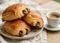 Golden flaky pain au chocolat pastries with melted chocolate batons visible at the ends, stacked on a plate
