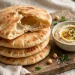 A stack of golden homemade pita breads, one torn open to show the hollow pocket inside, with hummus nearby