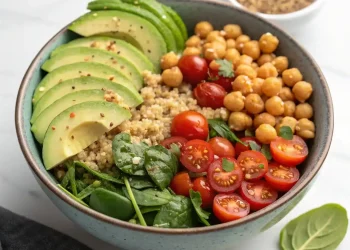 A high protein vegan salad bowl filled with sliced avocado, cooked chickpeas, halved cherry tomatoes, fresh spinach leaves, and quinoa arranged in sections. A small dish with seeds is visible in the background.