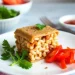 A square slice of baked beans sandwiched between two pieces of bread, garnished with parsley, sits on a white plate with sliced cherry tomatoes—a Quick Recipe from Jalbiteworldfood. Bowls of herbs and sauce are blurred in the background.