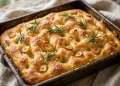 Golden focaccia with deep dimples, fresh rosemary, roasted garlic slices, and flaky salt in a pan