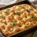 Golden focaccia with deep dimples, fresh rosemary, roasted garlic slices, and flaky salt in a pan