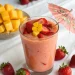 A glass of Strawberry Mango Smoothie topped with sliced strawberries, mango cubes, a yellow flower, and a decorative paper umbrella, surrounded by fresh strawberries and a bowl of mango cubes on a white surface.