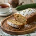 A sliced loaf of cooks zucchini bread dusted with powdered sugar sits on a wooden tray, with a cup of tea and a cucumber in the background.